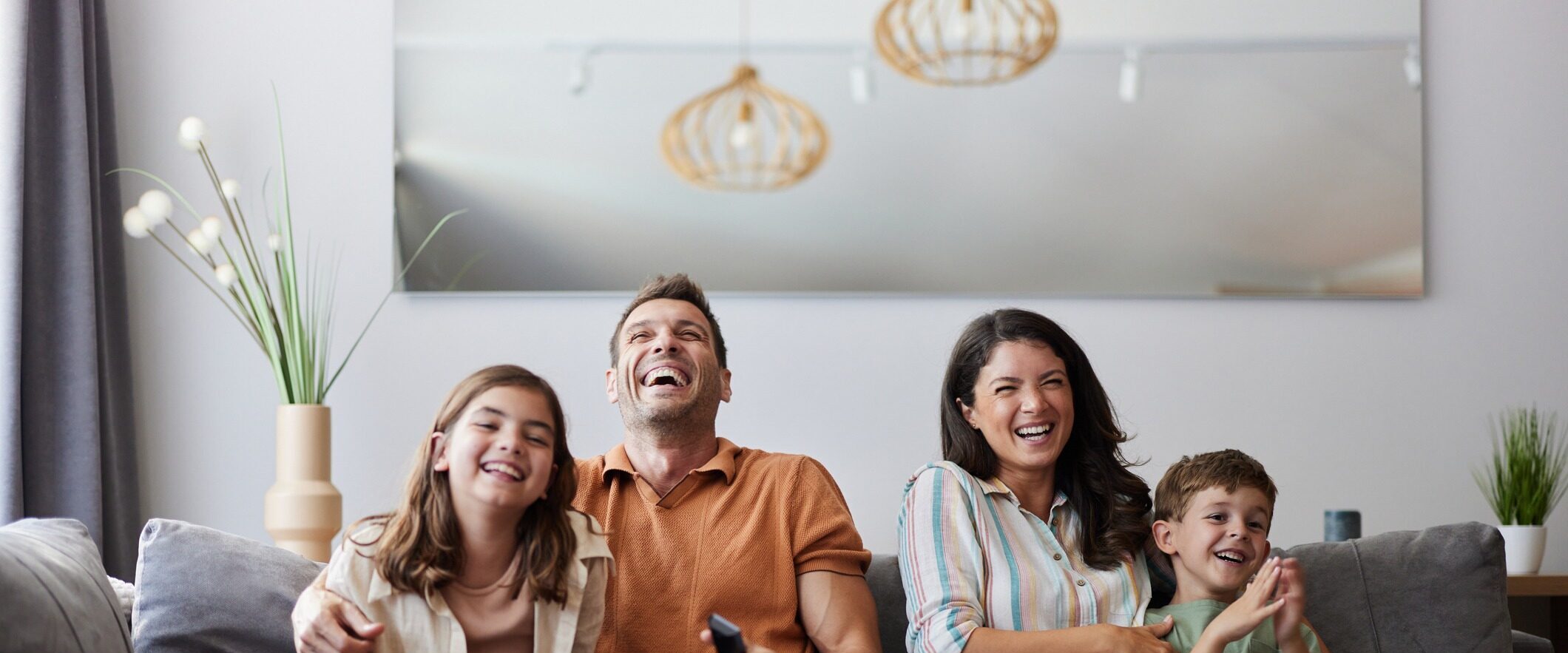 A family of four sits on a couch in a living room, all smiling and laughing. Two hanging lights and a mirror are visible in the background, along with a vase of tall grass on a side table.
